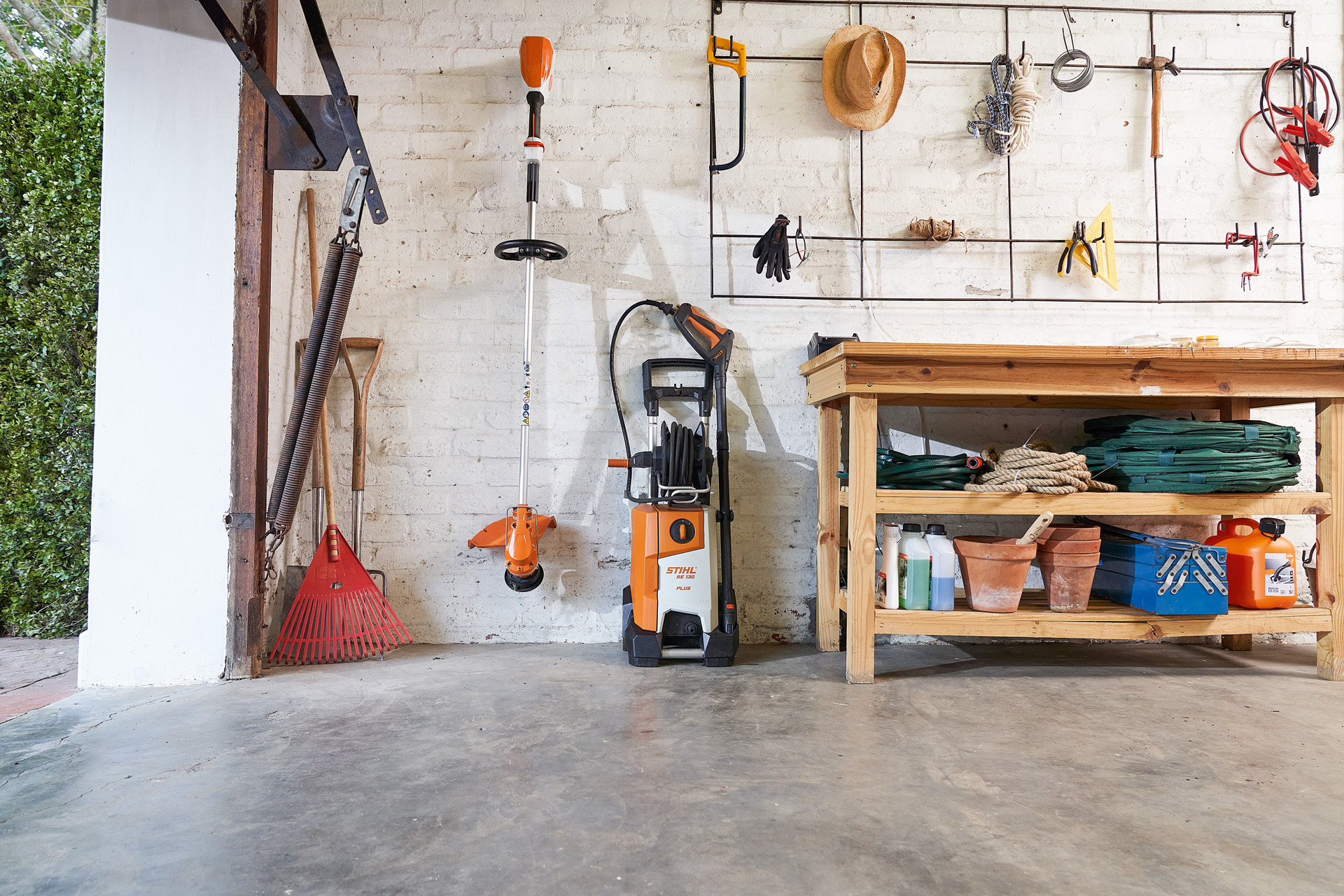 Una hidrolimpiadora STIHL RE 130 plus y una desbrozadora STIHL en un garaje junto a otras herramientas de jardín. 
