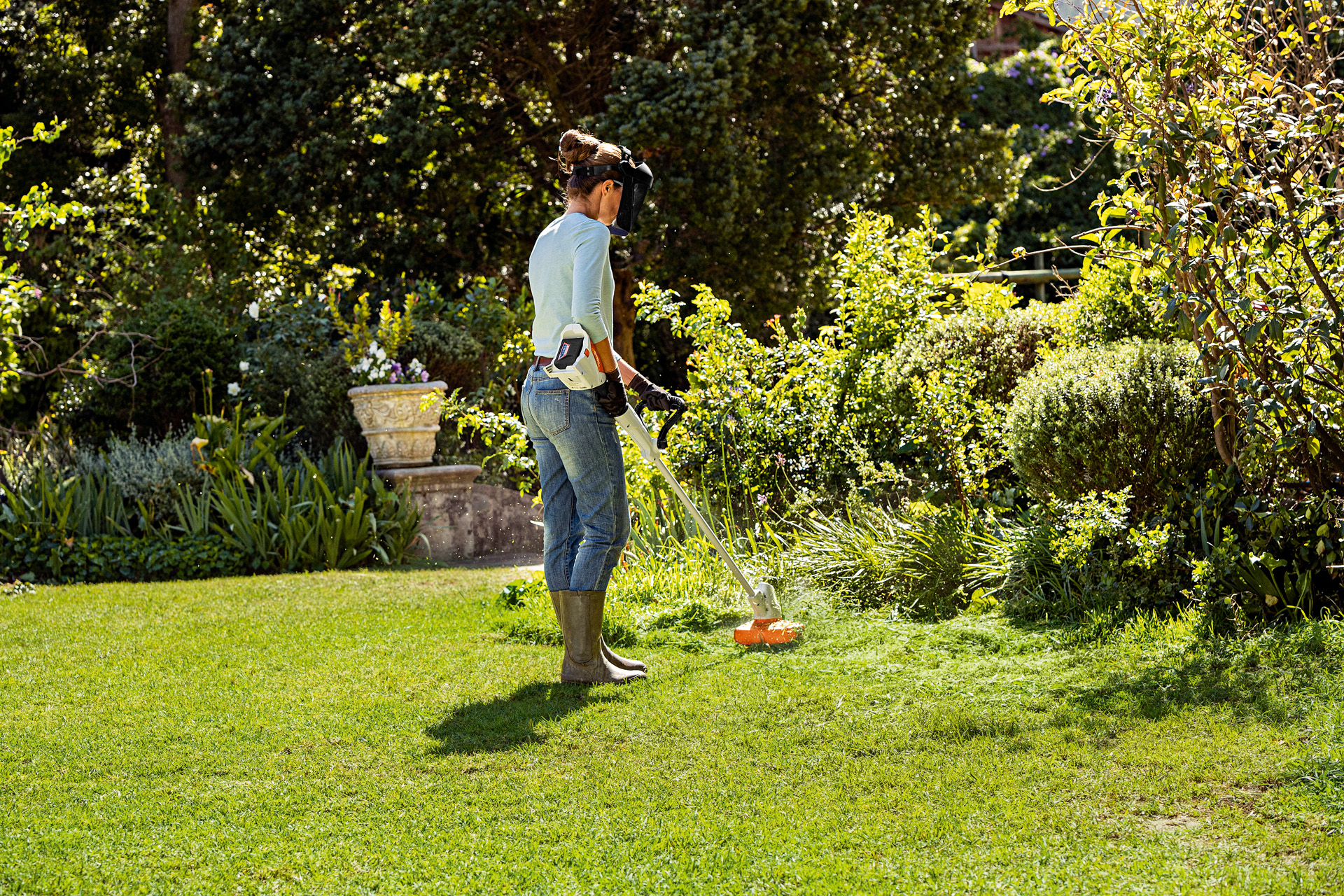 Una mujer con una pantalla protectora y guantes usando un cortabordes de batería STIHL FSA 56 en un jardín