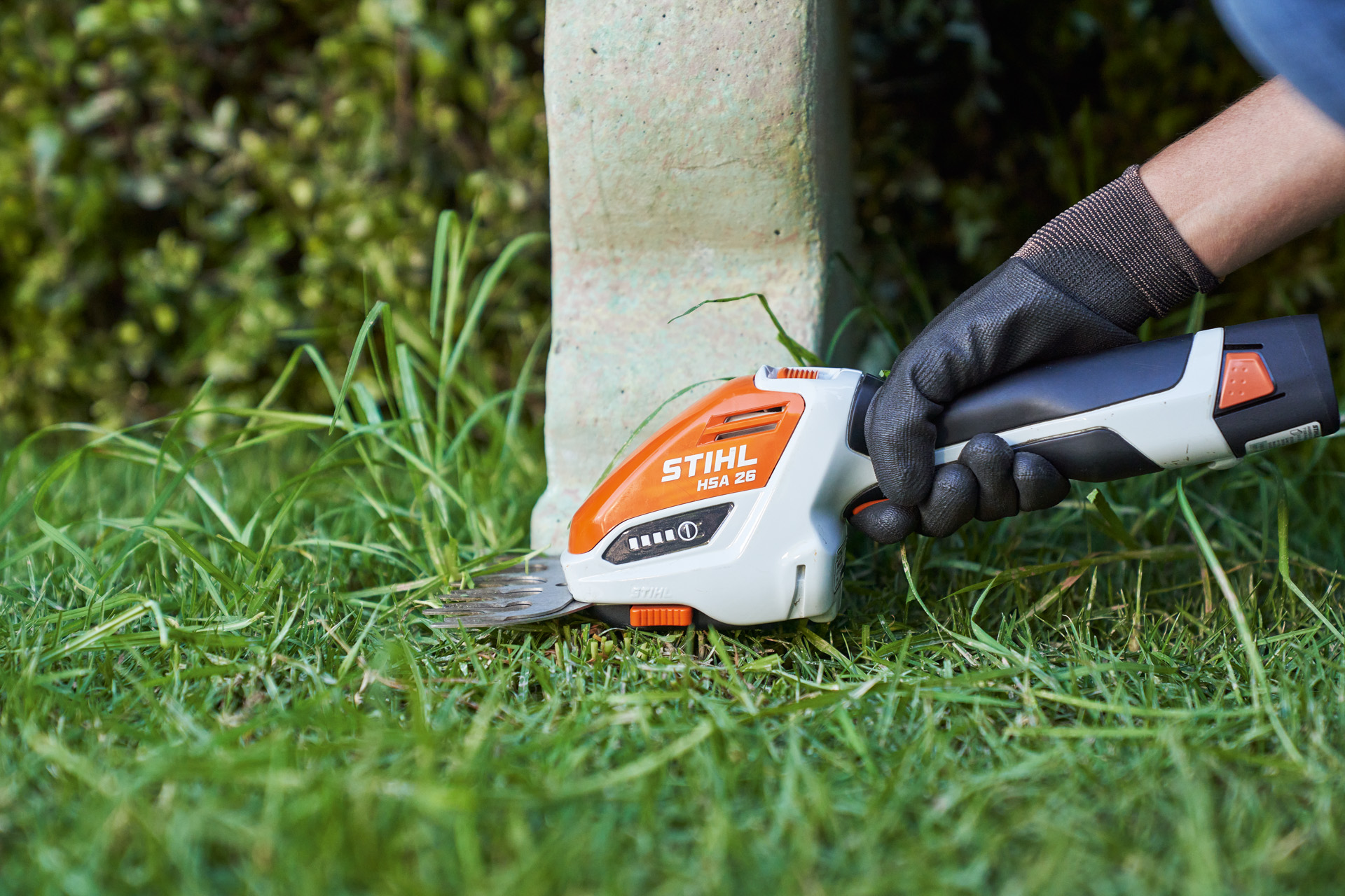 Una mano con guante de trabajo corta el borde del césped alrededor de un banco con el cortasetos de batería STIHL HSA 26