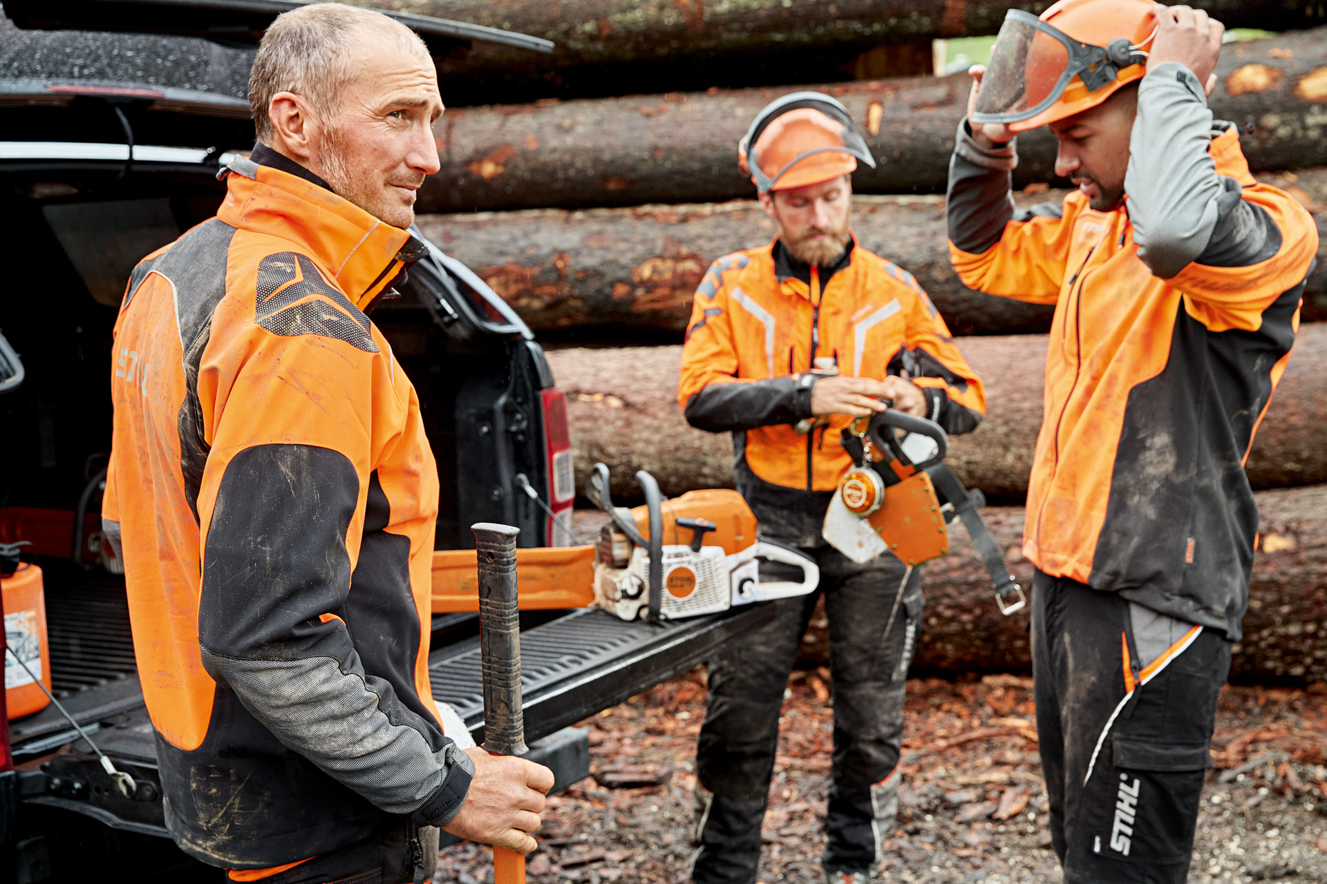 Tres hombres con ropa de protección STIHL se preparan para trabajar, con una motosierra STIHL junto a un vehículo