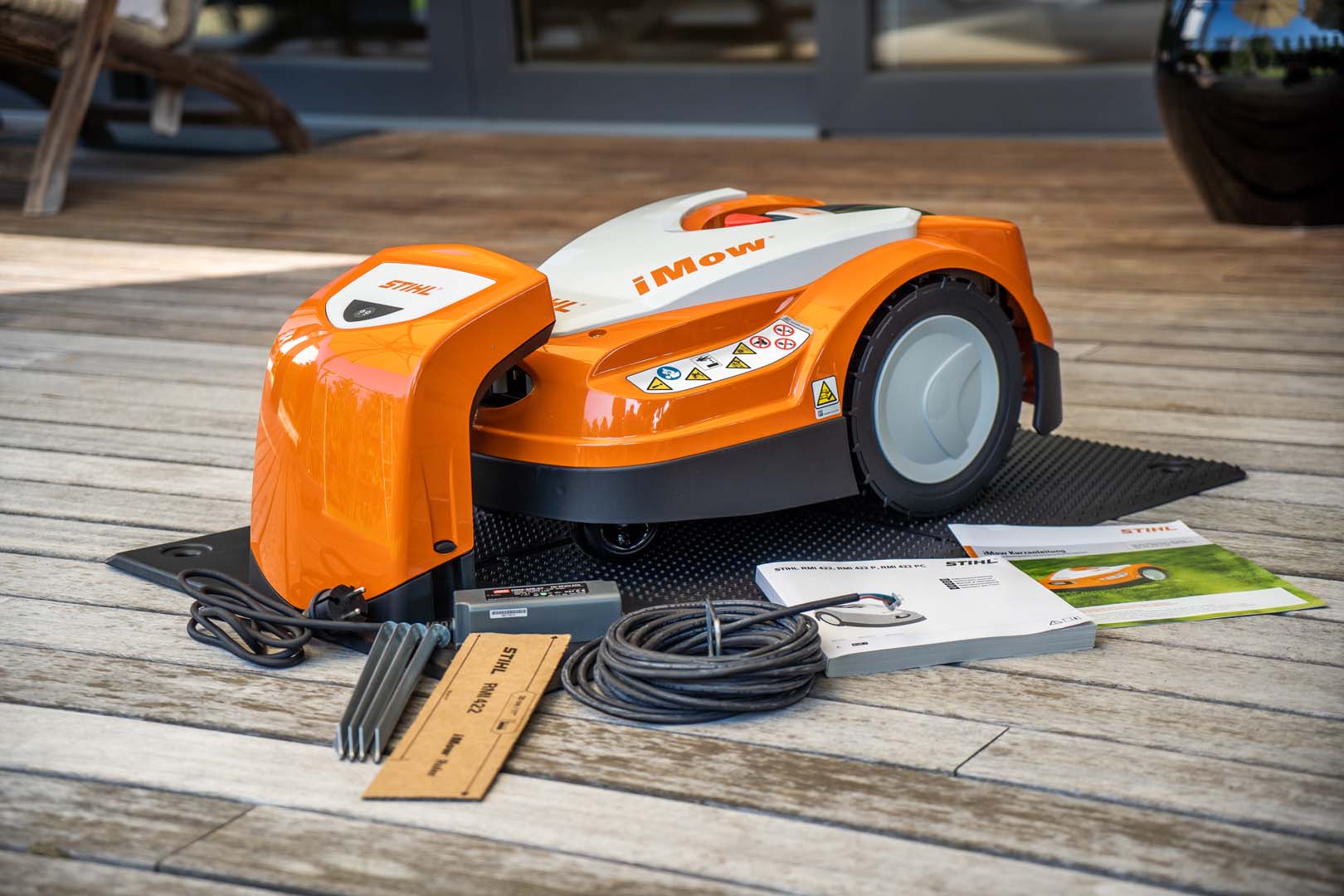 Un robot cortacésped STIHL iMOW® 422 en una terraza de madera con un cable y las instrucciones de funcionamiento