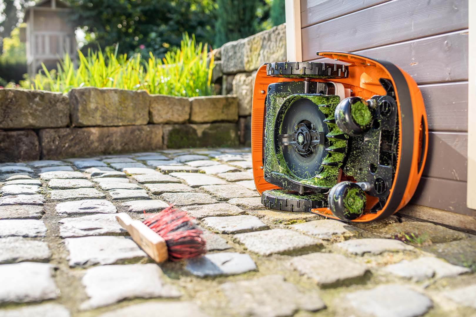 Un robot cortacésped STIHL iMOW® colocado sobre su lateral listo para la limpieza, con un cepillo cerca en el suelo
