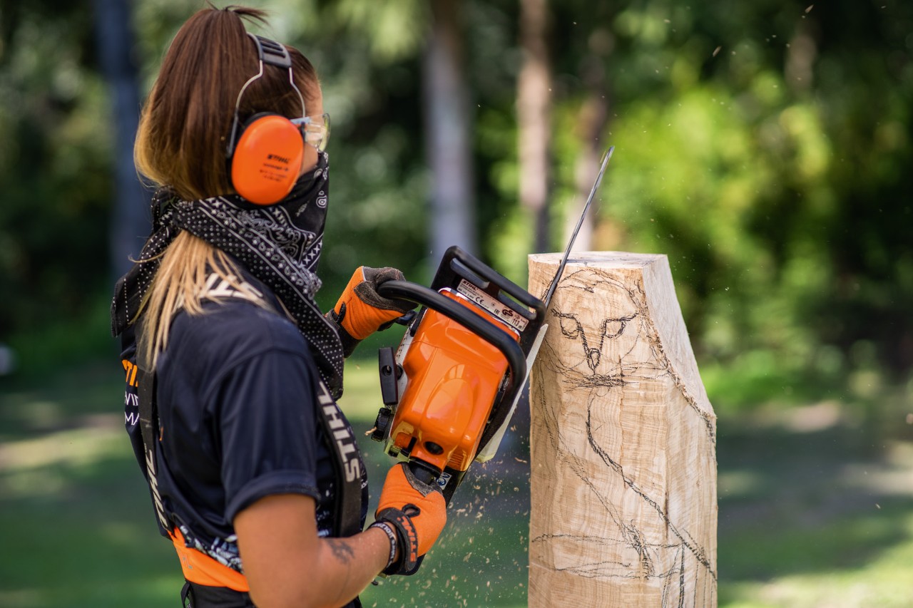 Mujer con protección auditiva, guantes y un pañuelo tapándole la boca mientras utiliza una motosierra STIHL para cortar a grandes rasgos un trozo de madera y empezar a tallar el búho.