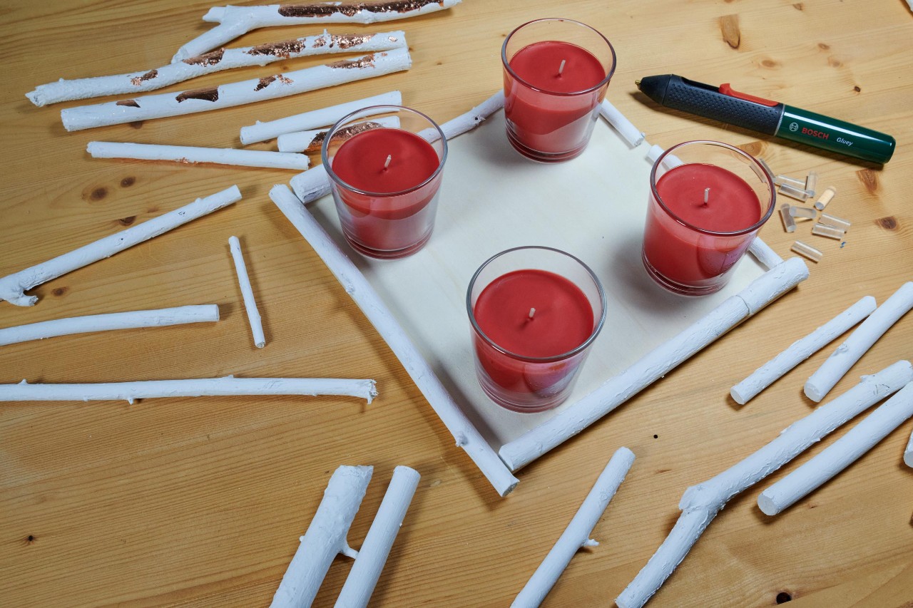 Crea tus coronas de Adviento de ramas secas: cuatro velas rojas en portavelas sobre una tabla de madera cuadrada rodeada de pequeñas ramas blancas