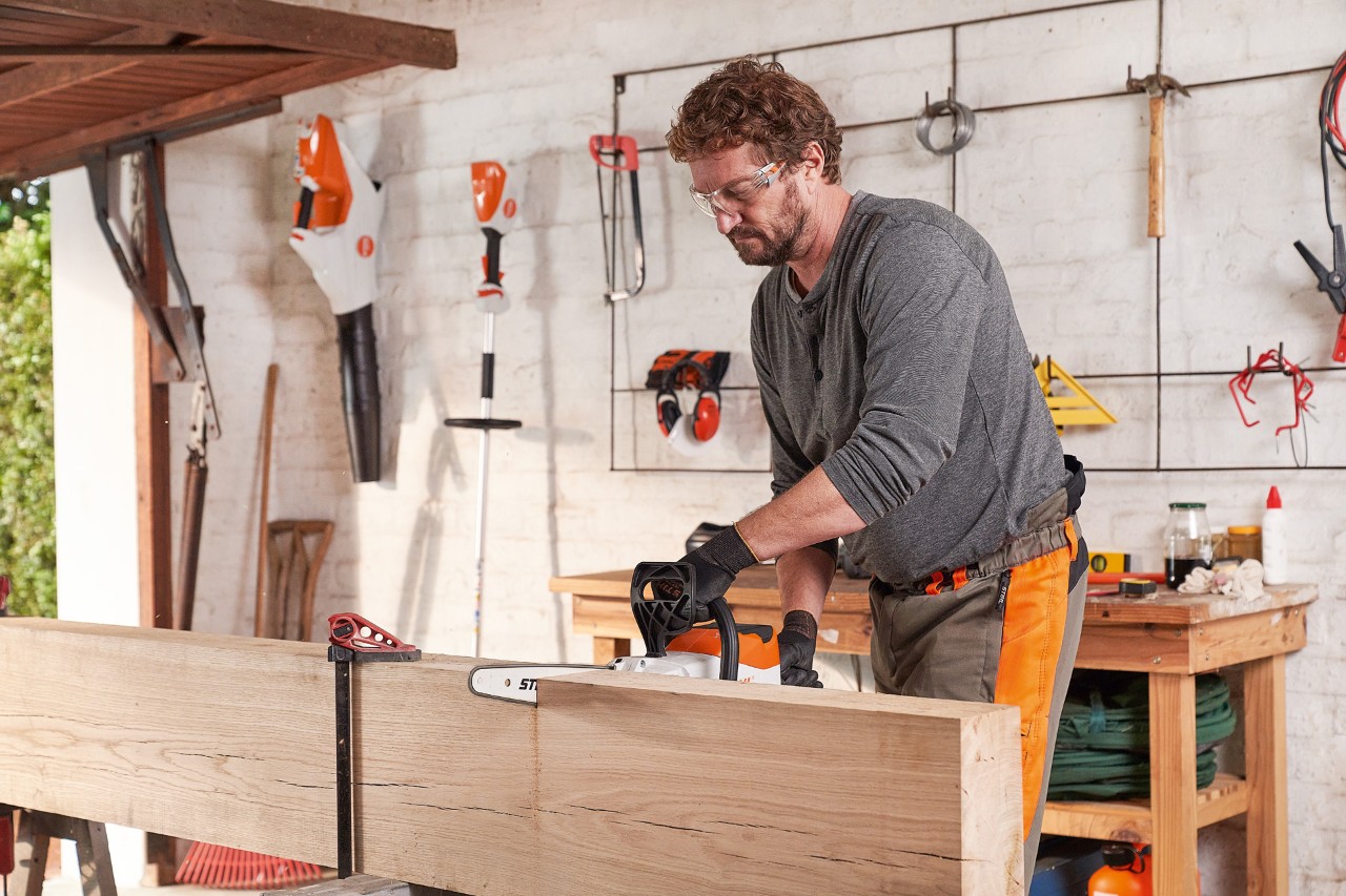 Un hombre con equipamiento de protección individual en el taller cortando un tablón con una motosierra de batería STIHL