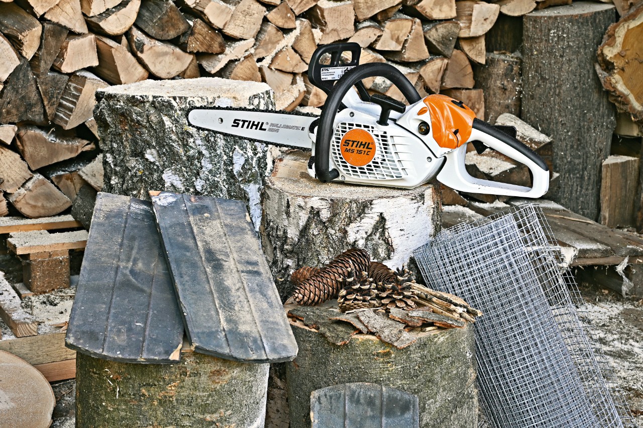 Una motosierra eléctrica STIHL MS 151 C, una malla de alambre, tejas, piñas y cortezas sobre tocones de árboles.