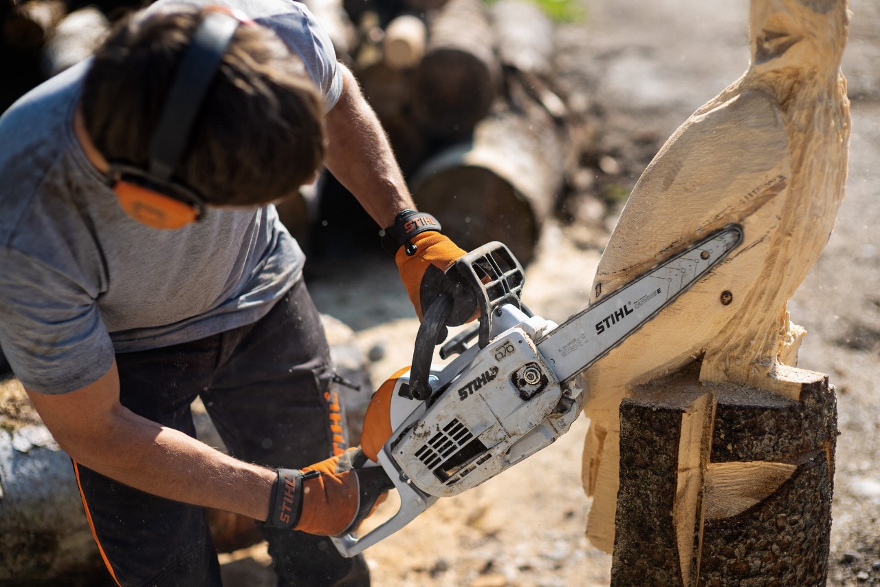Un hombre con protección auditiva utilizando una STIHL MS 193 C-E con accesorio de talla plumas en una escultura.