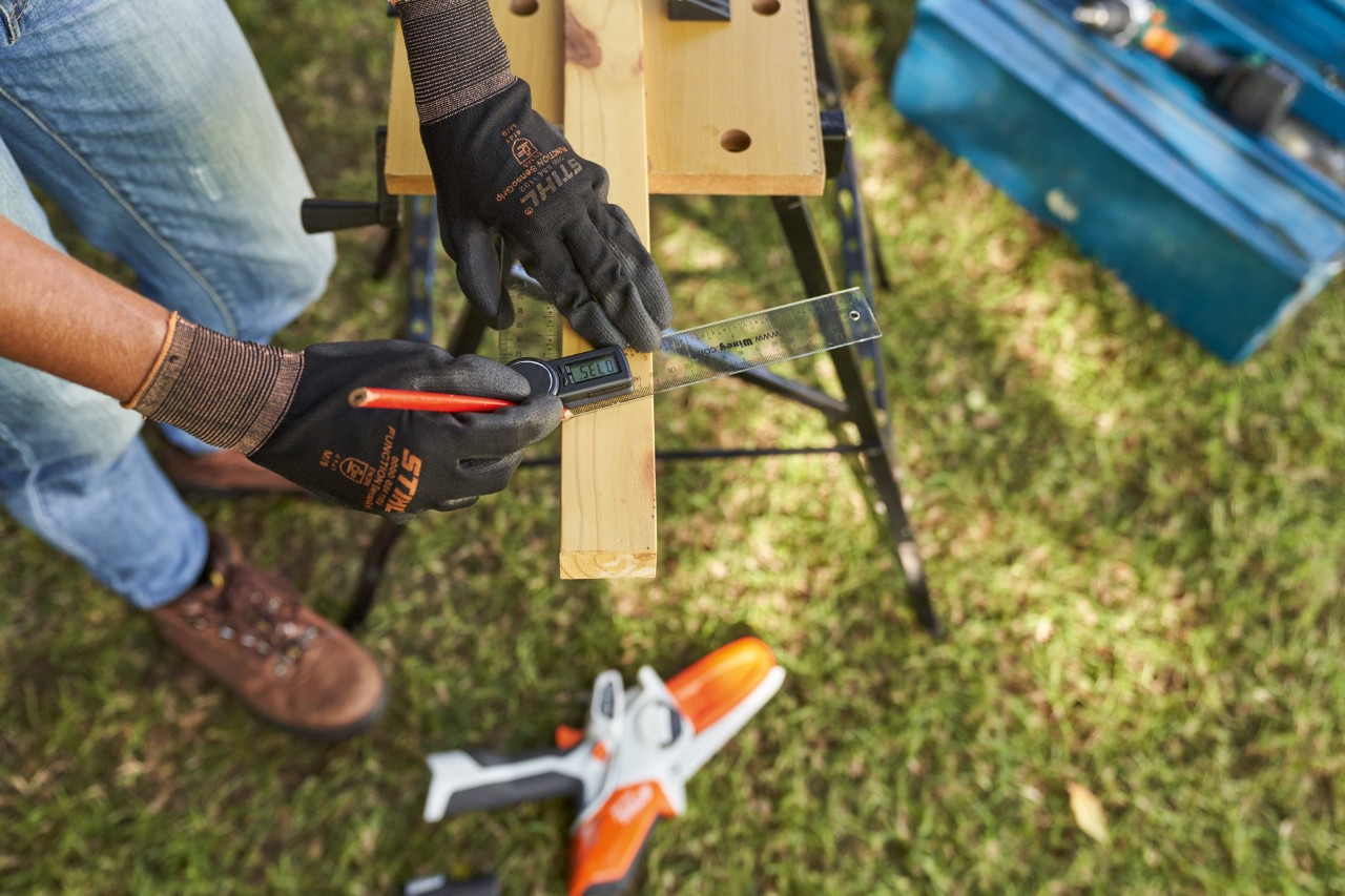 Una mujer con guantes de trabajo mide madera de construcción; abajo se puede ver la minisierra de batería STIHL GTA 26