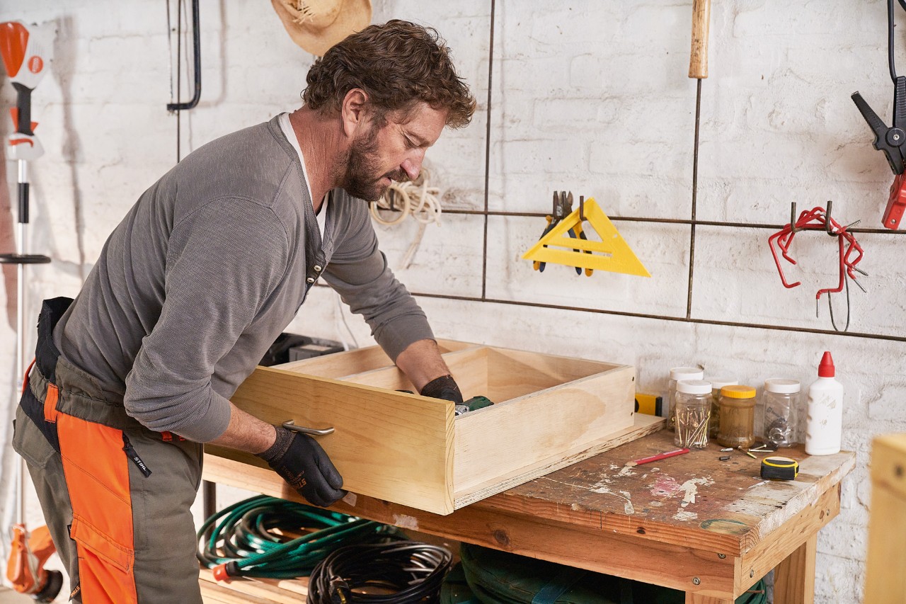 Un hombre fija un panel de pino a un cajón para construir él mismo el garaje para  cortacésped