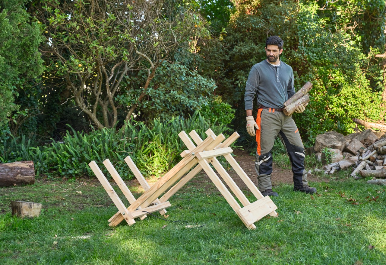 Un hombre en un jardín lleva troncos de madera hacia un caballete de madera construido por él mismo