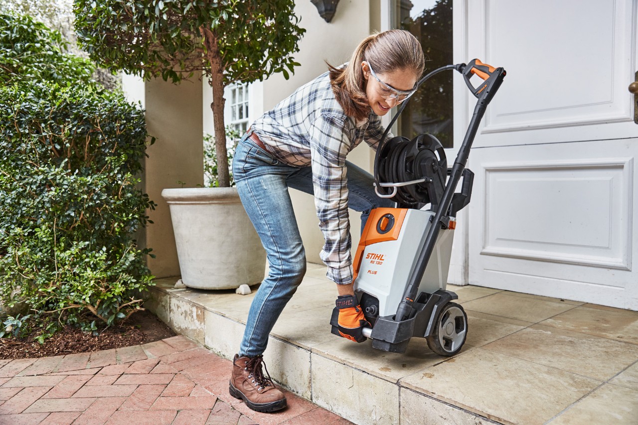 Una mujer levantando una hidrolimpiadora STIHL RE 130 Plus para sortear un escalón que lleva a un camino de ladrillos.