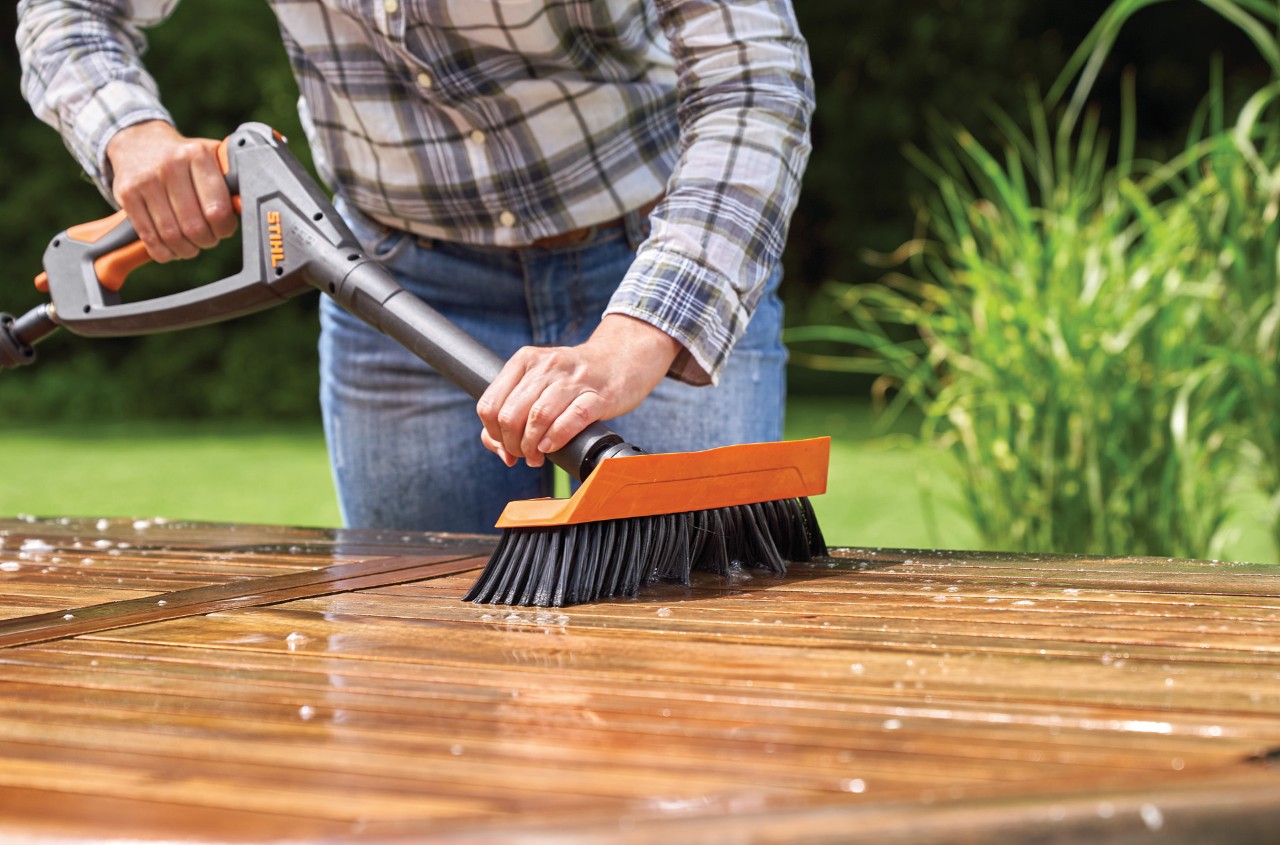 Una mujer hace un tratamiento para muebles de madera exterior con una hidrolimpiadora STIHL RE 130 Plus y un cepillo.