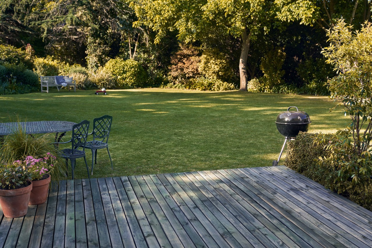 Terraza de madera frente a un jardín verde con barbacoa, sillas y macetas con plantas