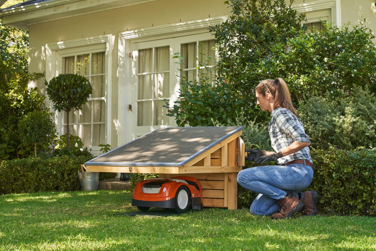 Una mujer arrodillada en el césped junto al garaje STIHL iMOW® abre el compartimento portaobjetos del robot cortacésped.