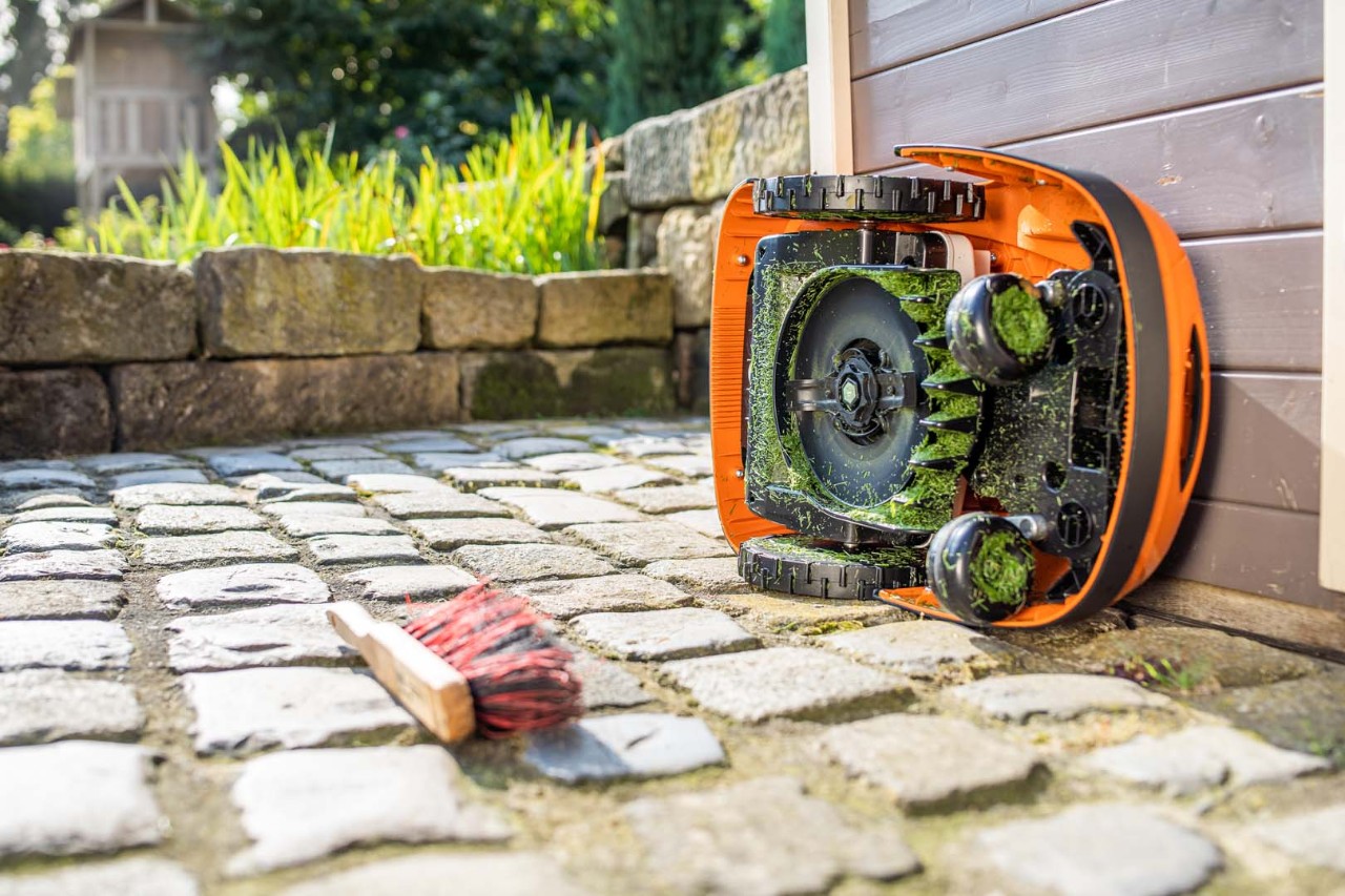 Un robot cortacésped STIHL iMOW® colocado sobre su lateral listo para la limpieza, con un cepillo cerca en el suelo