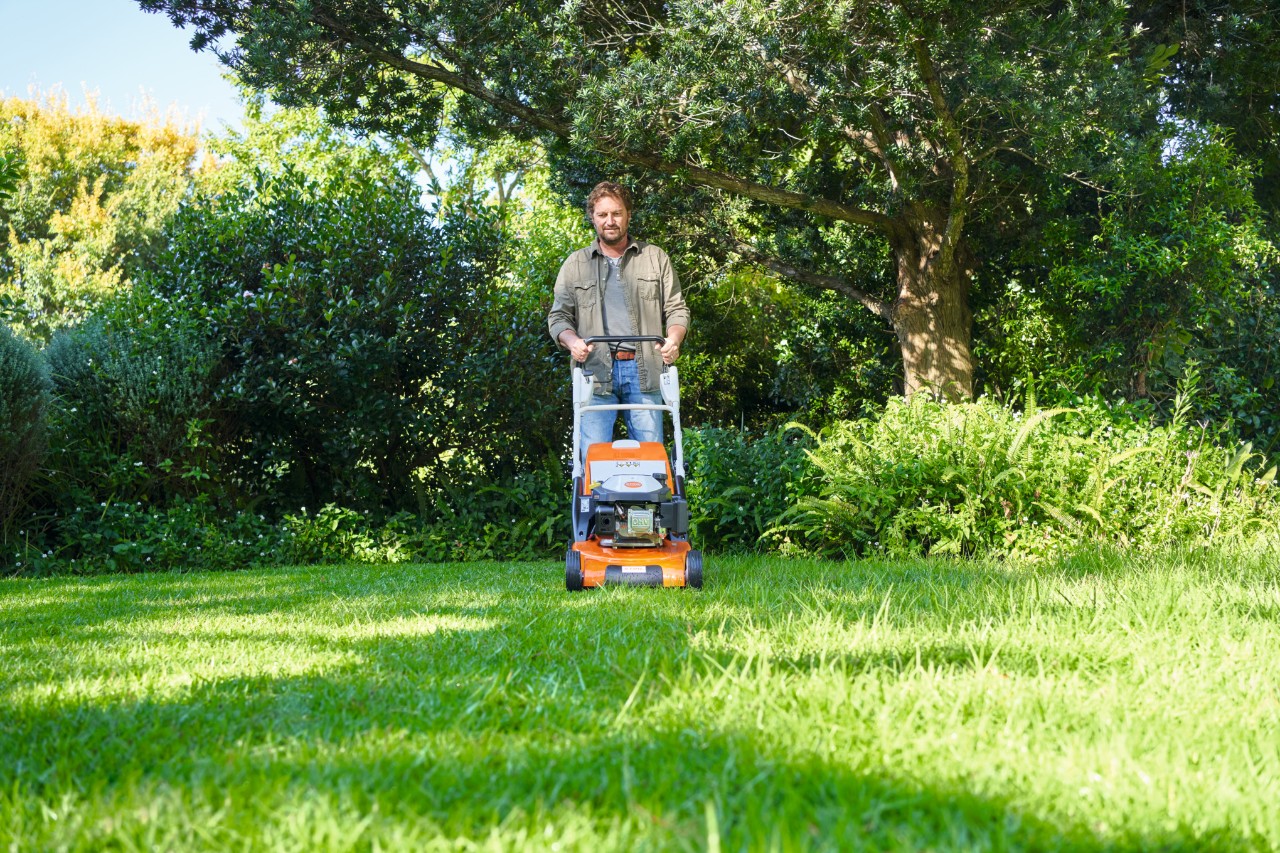 Un hombre corta un césped semisombreado con un cortacésped STIHL, con arbustos y árboles en segundo plano