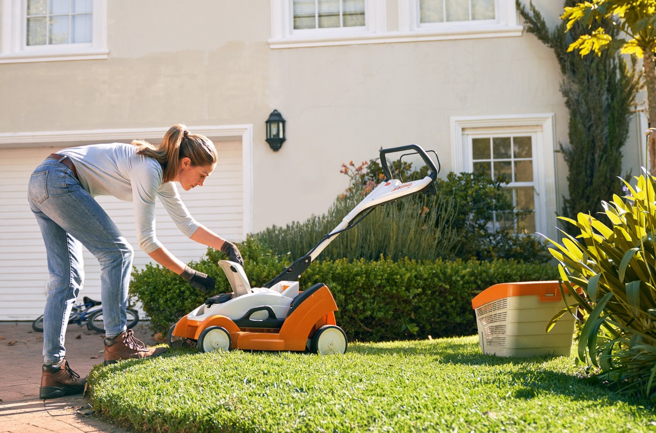 Una mujer abre la cubierta de un cortacésped de batería STIHL RMA 339 C delante de una casa blanca.