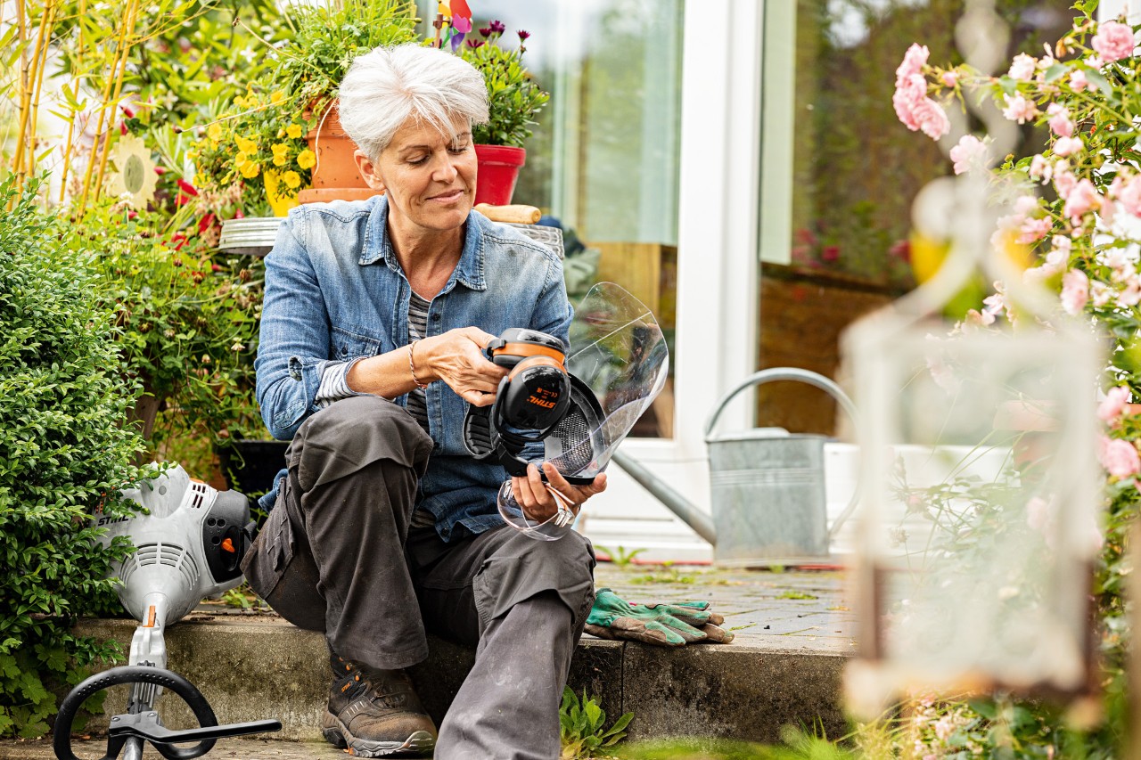 Una mujer está sentada entre plantas junto a una desbrozadora STIHL FS 38 observando el equipamiento de protección