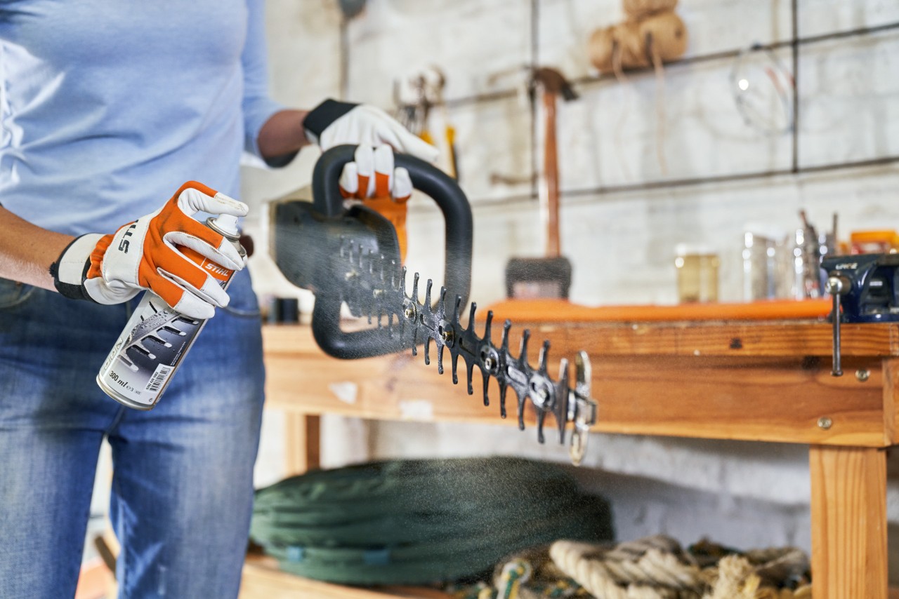 Una mujer con guantes de protección pulveriza un cortasetos de batería STIHL HSA 56 con spray de limpieza para el mantenimiento
