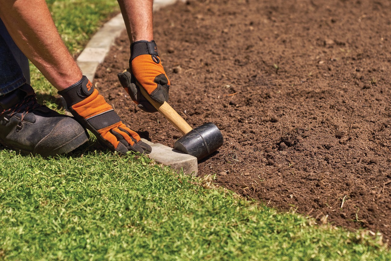 Una persona con guantes utiliza una maza para crear un borde de ladrillos para plantas perennes para parterres.