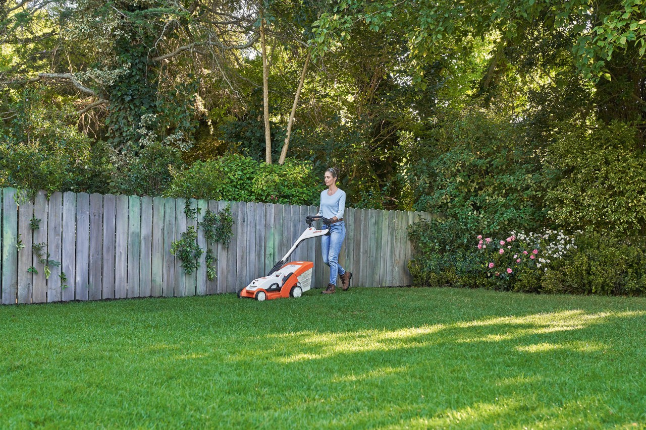 Una mujer corta el césped verde delante de una valla de madera con el cortacésped de batería STIHL RMA 339 C.