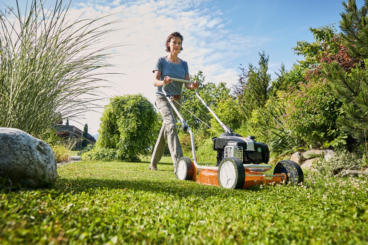 Una mujer utiliza un cortacésped para mulching STIHL RM 2 R de gasolina en un césped rodeado de arbustos y hierbas.