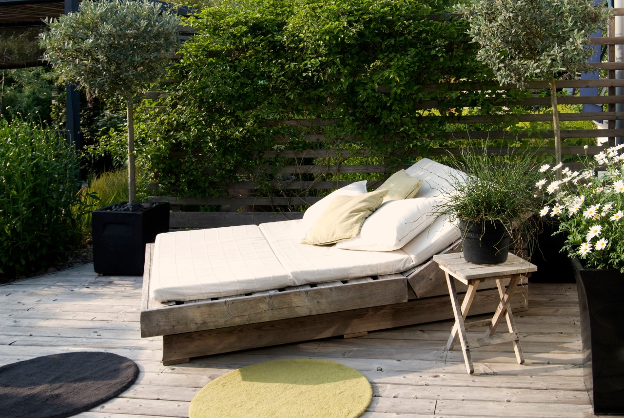Una terraza de madera con una tumbona con colchoneta y olivos pequeños al fondo