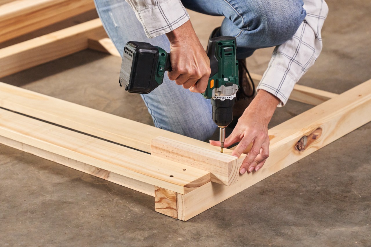 Primer plano de una persona atornillando con un destornillador eléctrico los listones de madera para la compostera.