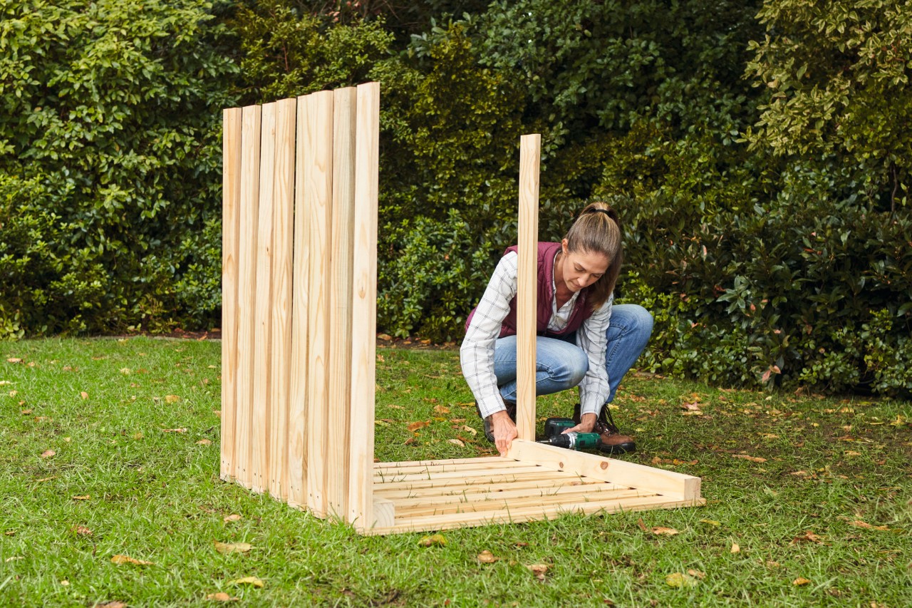 Una mujer fija los listones para las paredes laterales del compostador con un destornillador eléctrico en un jardín.