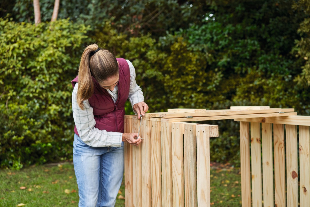 En un jardín, una mujer coloca ganchos en una compostera de construcción propia.