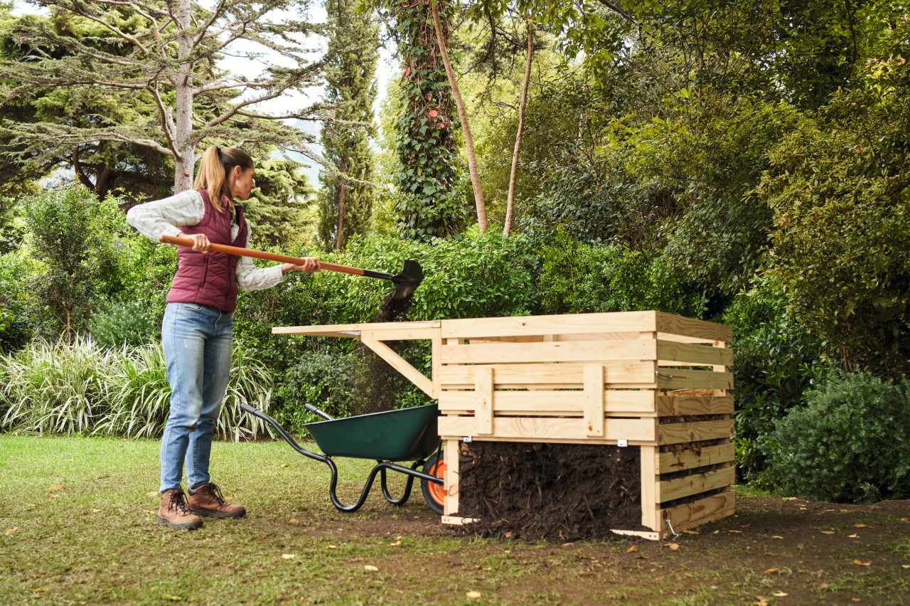 Una mujer en una parcela de jardín tamiza el compost en bruto junto a un compostador de madera de construcción propia