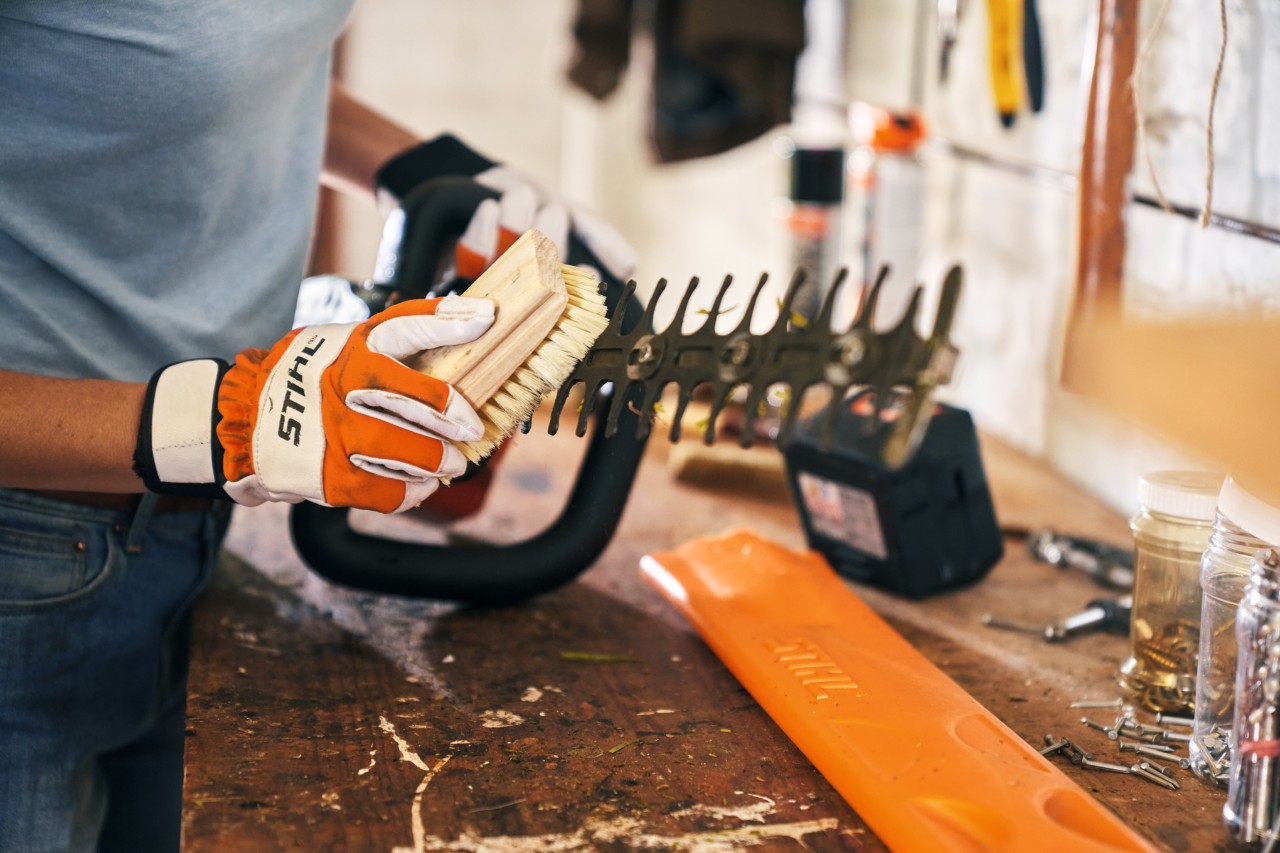 Una persona con guantes de protección limpia en un taller un cortasetos de altura con un cepillo 