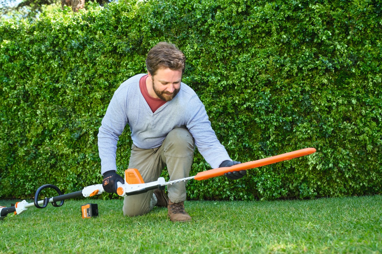 Un hombre delante de un seto coloca un cortasetos de altura de batería STIHL HLA 56 sobre el césped