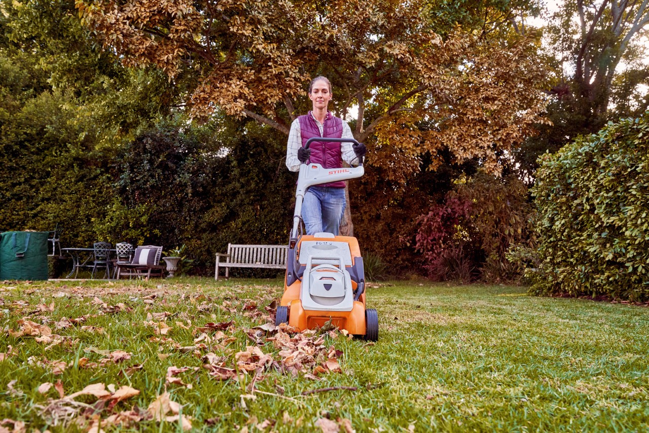 Una mujer corta un césped cubierto de hojas en un jardín de otoño con el cortacésped de batería STIHL RMA 339 C.