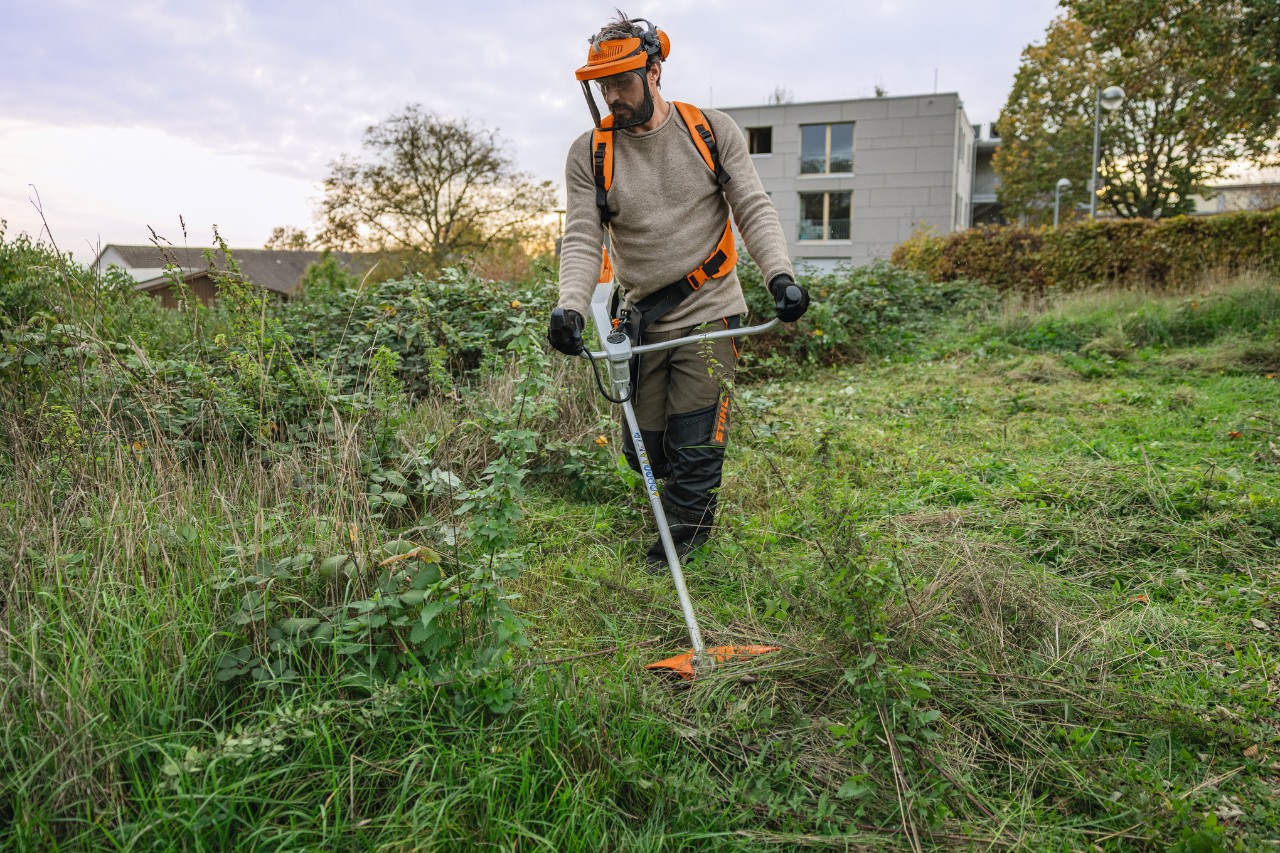 Un hombre con equipo de protección STIHL corta con una desbrozadora de batería STIHL FSA 200 una superficie verde de la que se han apoderado las malas hierbas