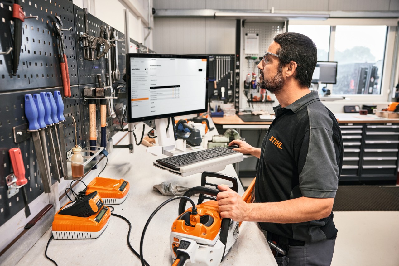 En un taller, un hombre se encuentra en primer plano manejando una computadora. Sobre la mesa frente a él hay una motosierra.