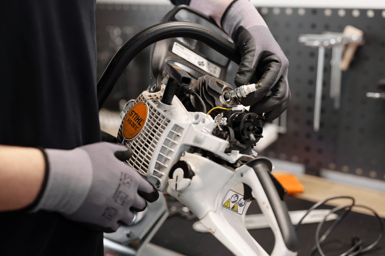 En un taller, una persona con guantes está de pie en primer plano, sosteniendo el motor de una motosierra y cambiando una bujía.