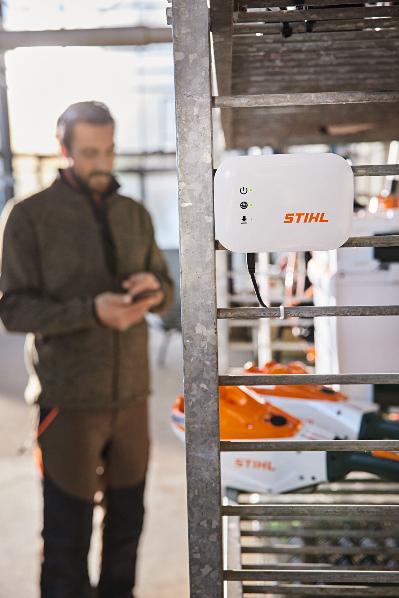 Una STIHL Connected Box cuelga de una estantería. Al fondo se ve a una persona que interactúa con su smartphone. 