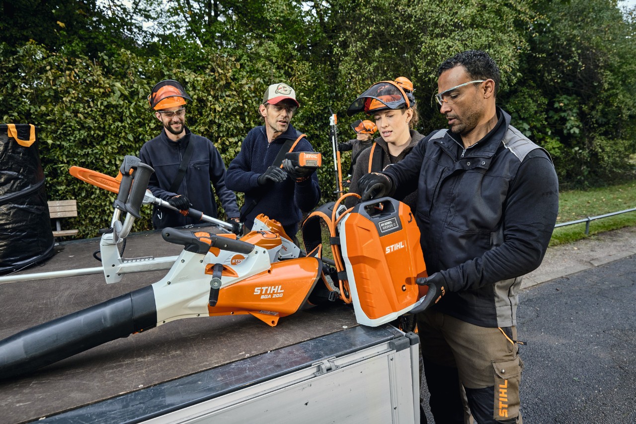 Tres hombres y una mujer con equipamiento de protección STIHL se encuentran en la zona de carga de un vehículo con máquinas STIHL, como el soplador de hojas de batería STIHL BGA 200 y la batería de mochila STIHL AR 2000 L 