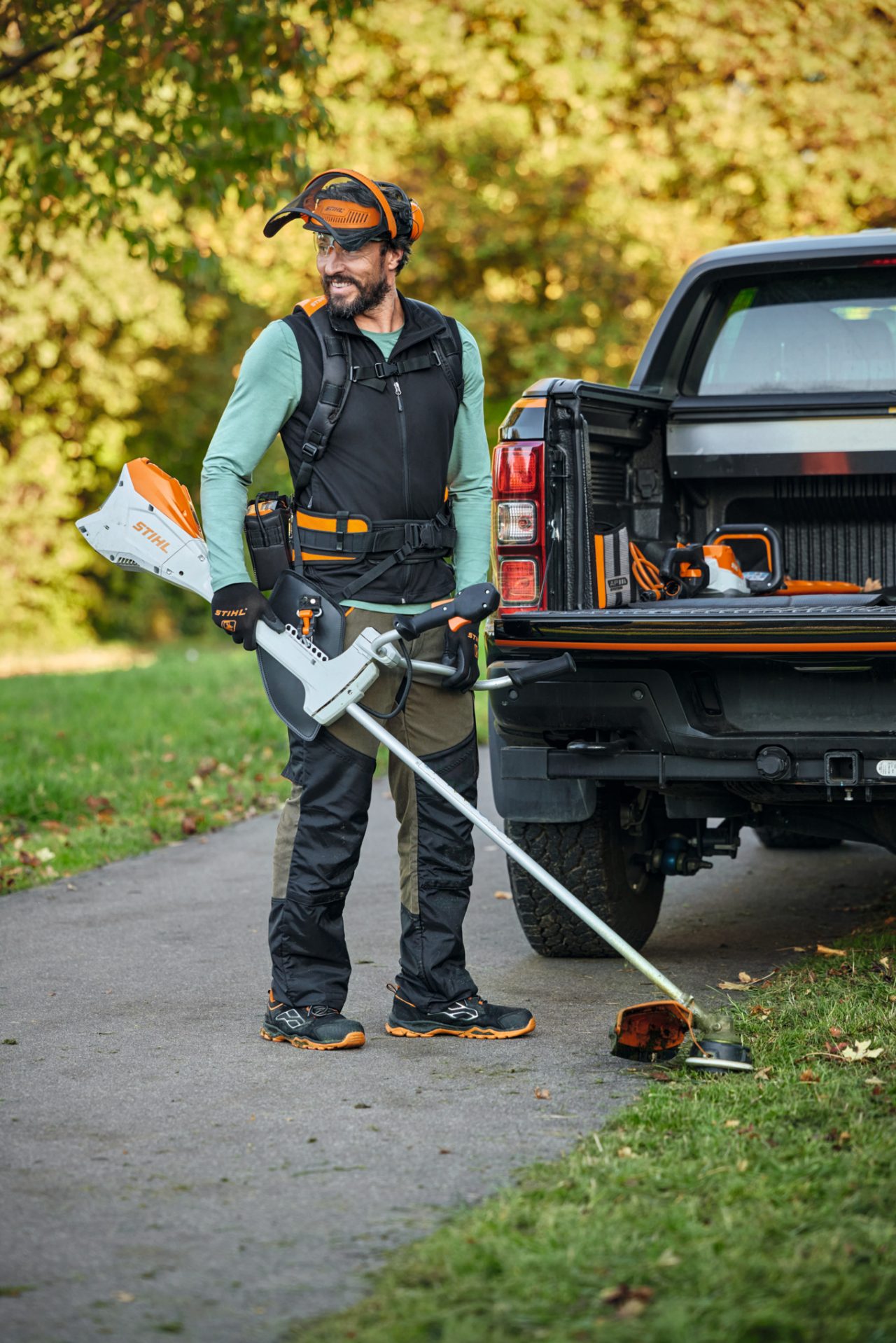 Un hombre con el sistema de arnés modular y una desbrozadora de batería STIHL FSA 200 delante de un coche