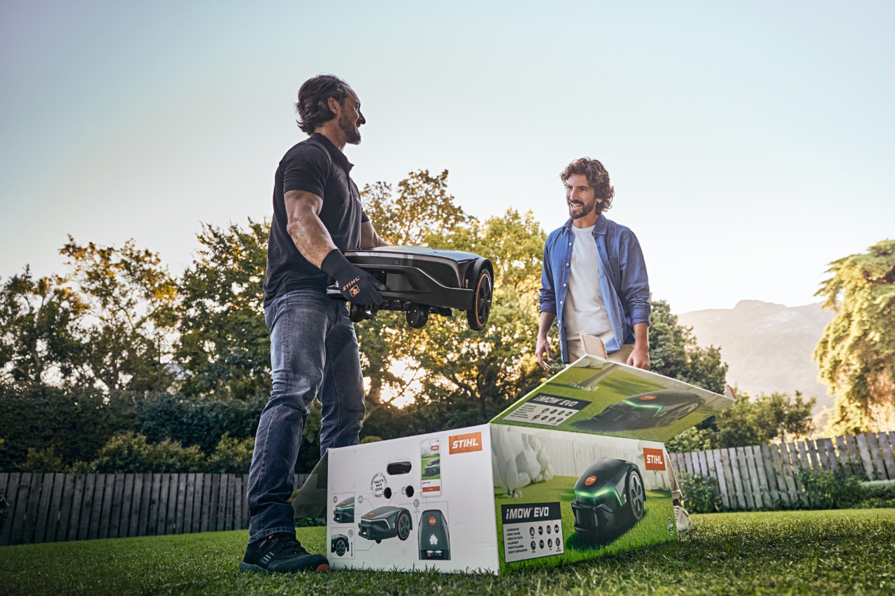 Una distribuidor especializado STIHL está en el jardín de un cliente con un iMOW® EVO en la mano y la caja de cartón iMOW® delante para empezar la instalación