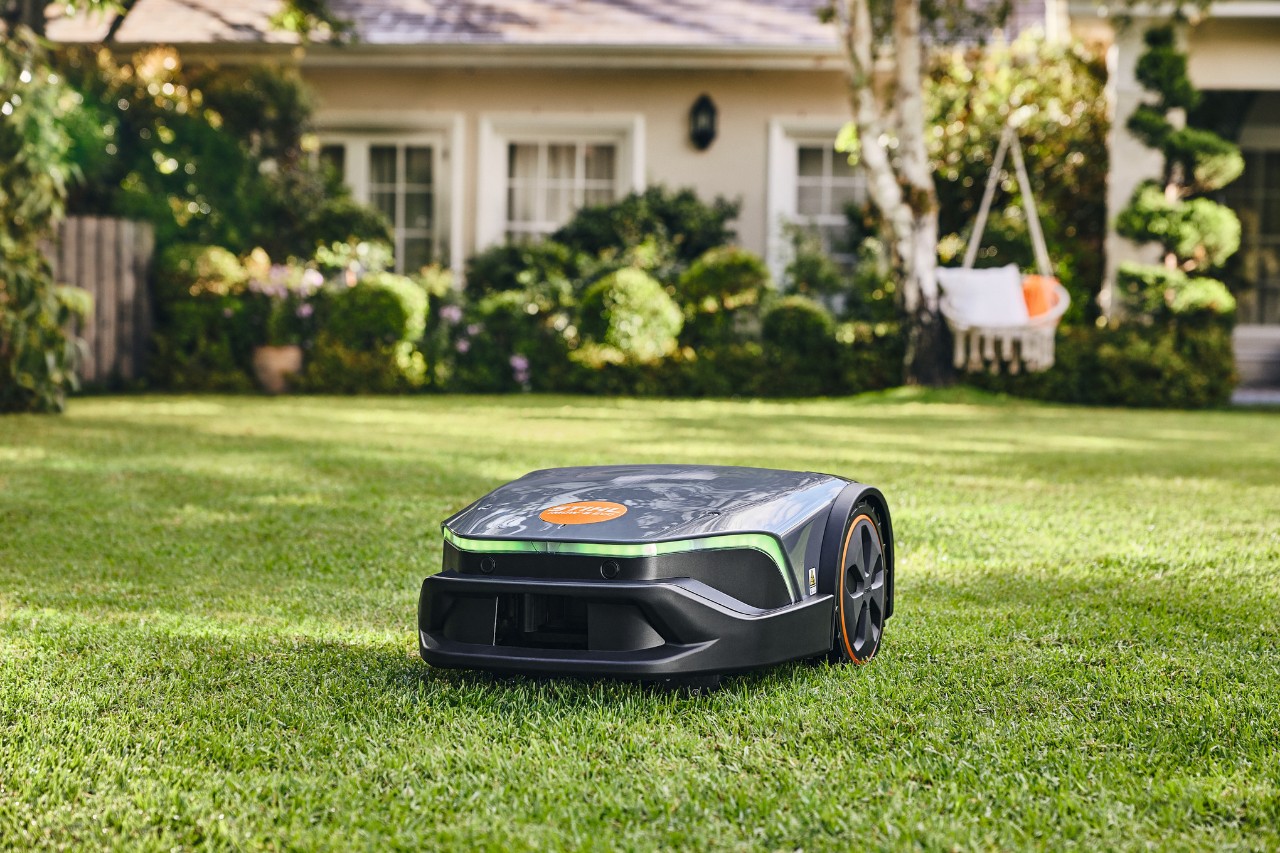 Un iMOW® Evo corta un césped bien cuidado en el jardín delante de una casa y una hamaca.